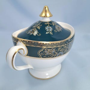 May include: A white porcelain sugar bowl with a lid, featuring a deep teal band decorated with gold floral patterns and a geometric border. The lid is topped with a gold finial, and the handle and base are accented with gold trim.