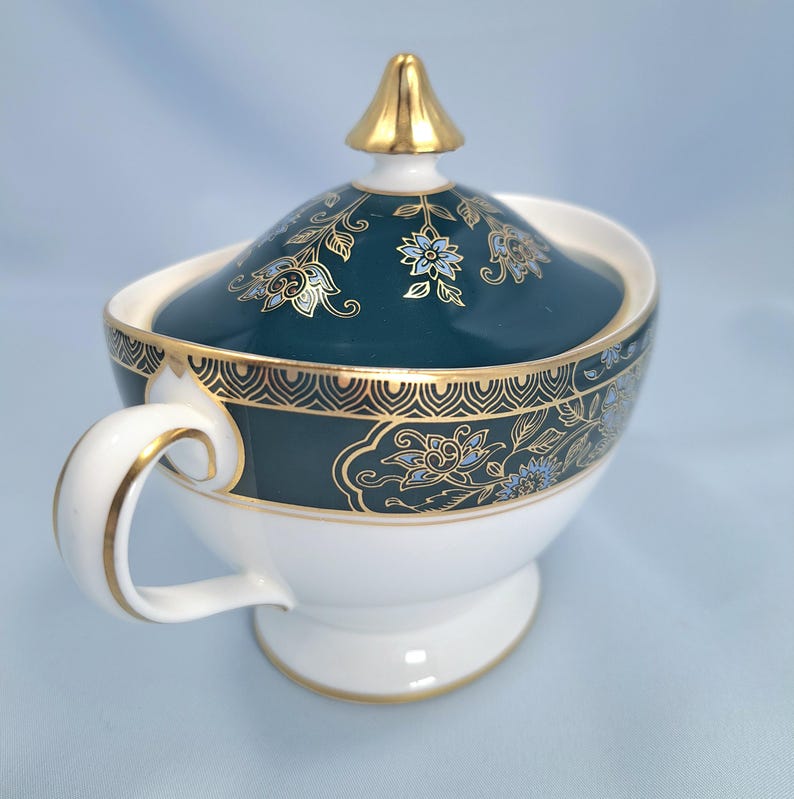 May include: A white porcelain sugar bowl with a lid, featuring a deep teal band decorated with gold floral patterns and a geometric border. The lid is topped with a gold finial, and the handle and base are accented with gold trim.