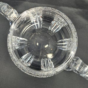 Op de afbeelding: Een ronde kom van helder glas met twee handvatten aan de zijkanten en een decoratief geslepen patroon. De buitenrand en de basis vertonen complexe geometrische facetten, terwijl de binnenwanden glad zijn met verticale accenten.
