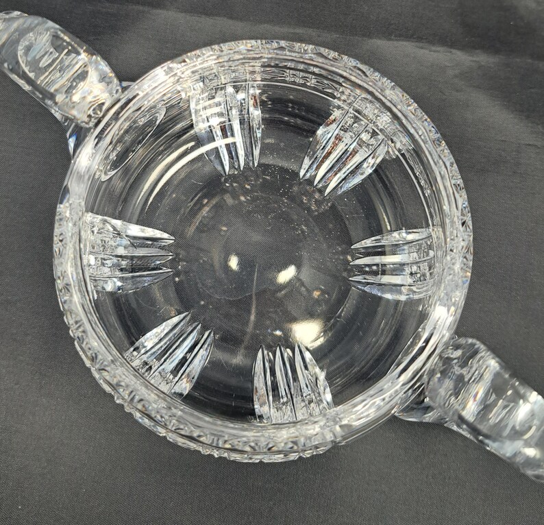 Op de afbeelding: Een ronde kom van helder glas met twee handvatten aan de zijkanten en een decoratief geslepen patroon. De buitenrand en de basis vertonen complexe geometrische facetten, terwijl de binnenwanden glad zijn met verticale accenten.