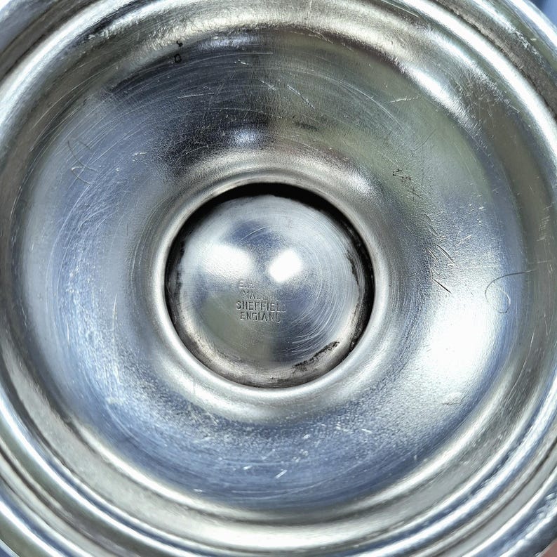 K&ouml;nnte beinhalten: Nahaufnahme eines silberfarbenen Metallobjekts, m&ouml;glicherweise ein Kerzenst&auml;nderfu&szlig;. Der Fu&szlig; hat eine abgerundete, konkave Form mit konzentrischen Ringen und einem zentralen, kugelf&ouml;rmigen Detail. Der Text "SHEFFIELD ENGLAND" ist sichtbar.