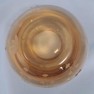 May include: A round, amber-colored glass bowl viewed from directly above. The bowl features a smooth, translucent surface with etched floral and botanical patterns around the outer rim, showcasing delicate stems and circular petal designs.