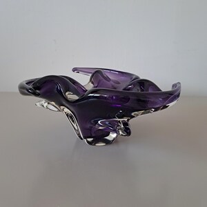 K&ouml;nnte beinhalten: Eine tiefviolette Glasschale mit unregelm&auml;&szlig;iger, organischer Form, flie&szlig;enden, geschwungenen Kanten und einem klaren Glasboden. Die Schale hat eine glatte, reflektierende Oberfl&auml;che mit verschiedenen Violett- und Dunkellila-T&ouml;nen im Glas.