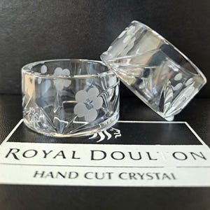 May include: Two clear crystal napkin rings featuring a delicate etched floral pattern and hand-cut geometric facets. The items are displayed on a black and white box labelled Royal Doulton Hand Cut Crystal, showcasing the brand and craftsmanship.