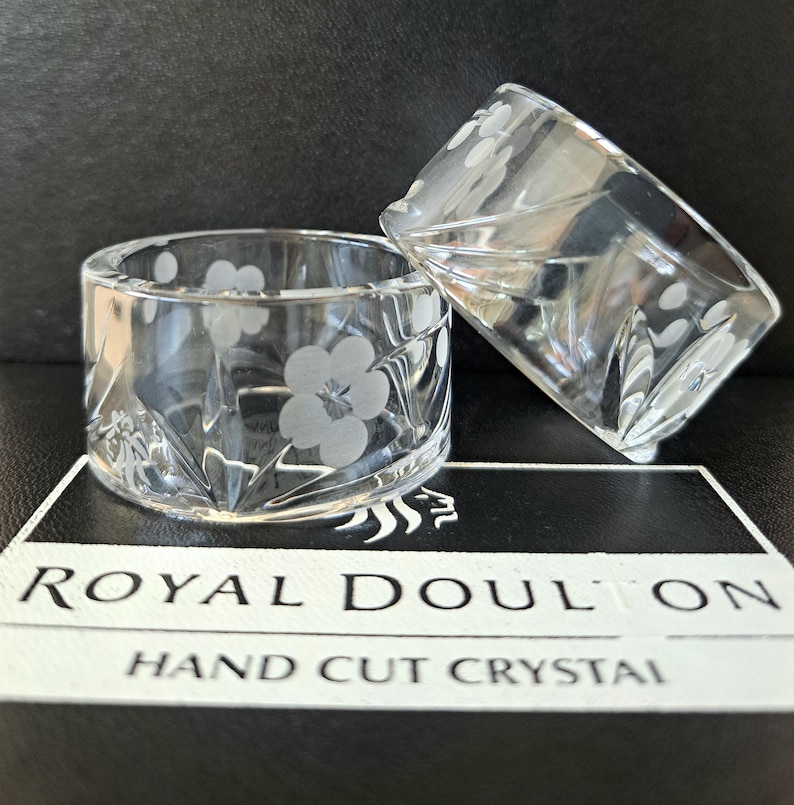 May include: Two clear crystal napkin rings featuring a delicate etched floral pattern and hand-cut geometric facets. The items are displayed on a black and white box labelled Royal Doulton Hand Cut Crystal, showcasing the brand and craftsmanship.
