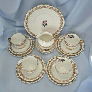 May include: A classic tea set comprising a large plate, a milk jug, a sugar bowl, and four teacups with saucers. The set is white with gold detailing and a pink rose design. The plates have a decorative, scalloped edge.