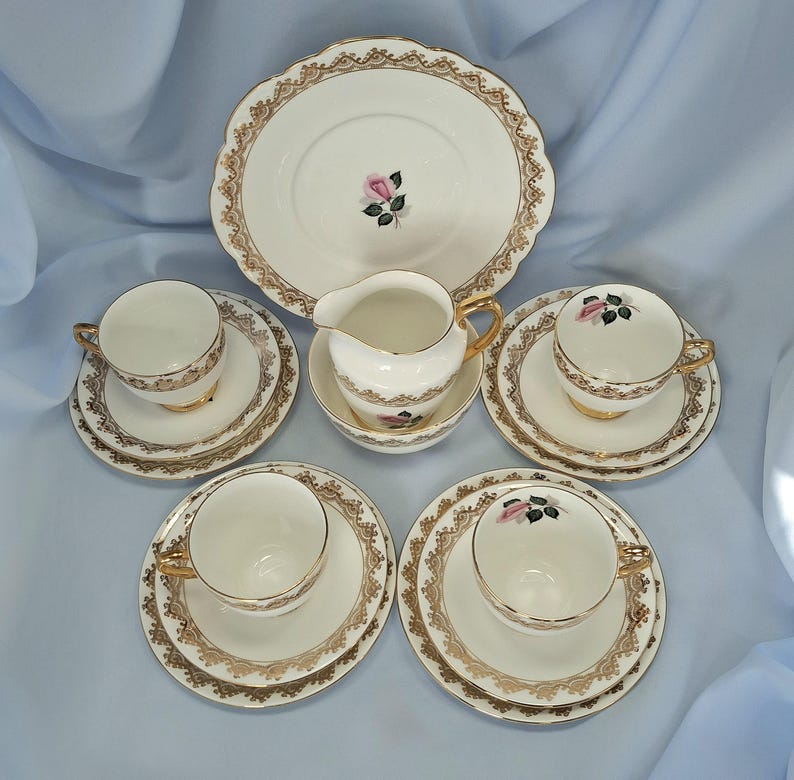 May include: A classic tea set comprising a large plate, a milk jug, a sugar bowl, and four teacups with saucers. The set is white with gold detailing and a pink rose design. The plates have a decorative, scalloped edge.