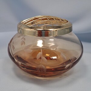 May include: A round, amber-colored glass bowl featuring a delicate etched leaf and floral pattern on its exterior. The bowl is topped with a removable, polished gold-tone metal grid lid designed for arranging flowers, creating a functional centerpiece.