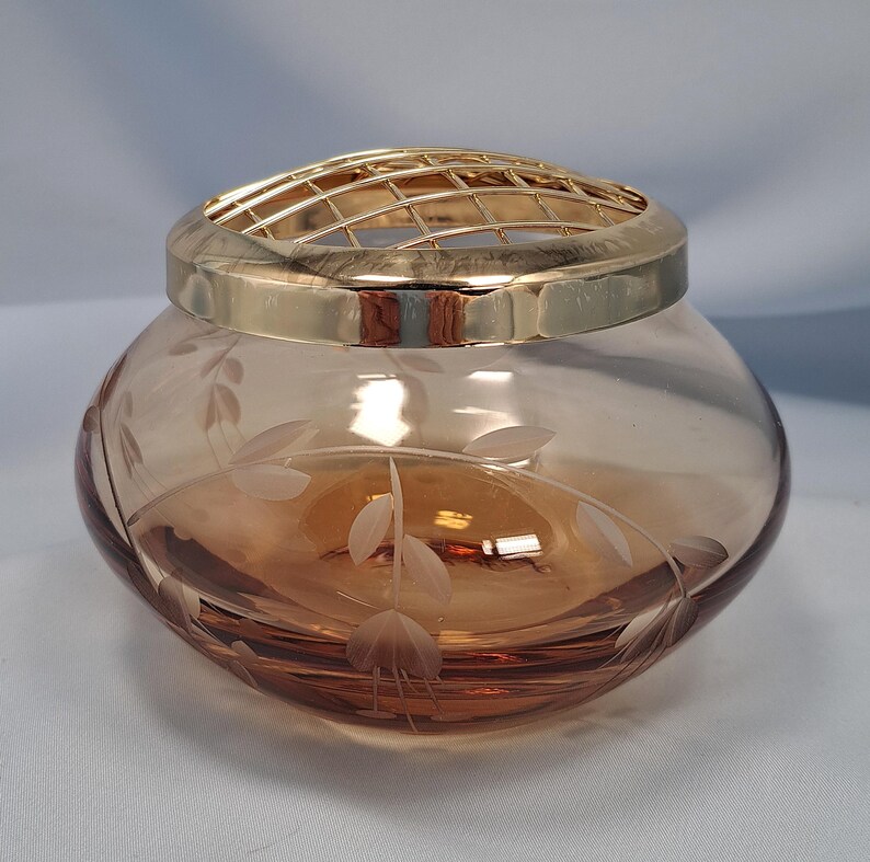 May include: A round, amber-colored glass bowl featuring a delicate etched leaf and floral pattern on its exterior. The bowl is topped with a removable, polished gold-tone metal grid lid designed for arranging flowers, creating a functional centerpiece.