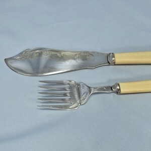 May include: Silver butter knife and fork set with ivory-colored handles. The knife has a curved blade with decorative etching. The fork has four tines and a matching handle. The set is displayed on a light blue surface.