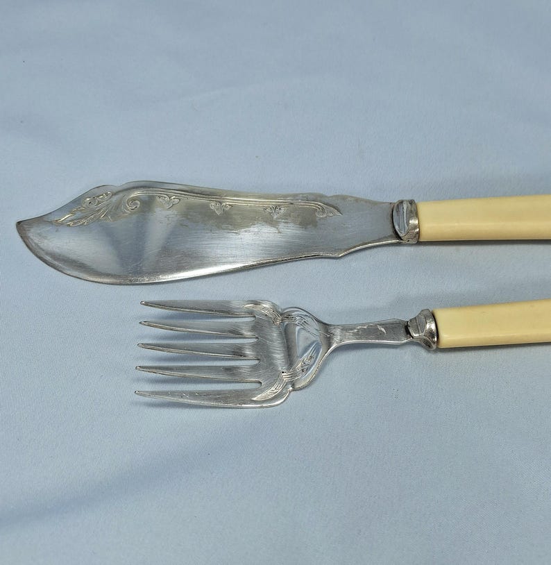 May include: Silver butter knife and fork set with ivory-colored handles. The knife has a curved blade with decorative etching. The fork has four tines and a matching handle. The set is displayed on a light blue surface.