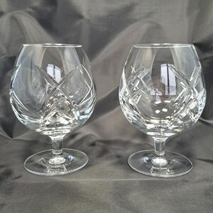Puede incluir: Dos copas de brandy de cristal transparente con un patr&oacute;n de corte profundo y complejo de l&iacute;neas cruzadas y estrellas. Cada copa tiene un cuenco ancho y redondeado que se estrecha hacia el borde, sobre un tallo corto y una base circular.