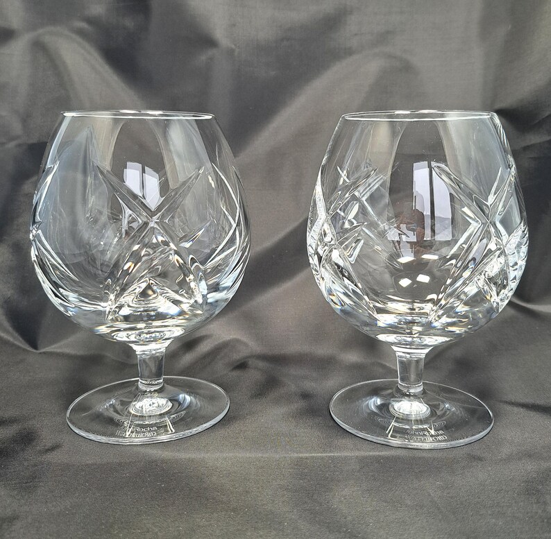 Puede incluir: Dos copas de brandy de cristal transparente con un patr&oacute;n de corte profundo y complejo de l&iacute;neas cruzadas y estrellas. Cada copa tiene un cuenco ancho y redondeado que se estrecha hacia el borde, sobre un tallo corto y una base circular.