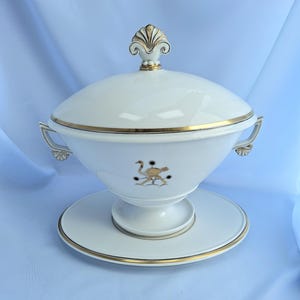 May include: A white ceramic soup tureen with a lid, handles, and a matching base. The tureen is decorated with gold trim and a stylized ostrich design. The lid is topped with a gold finial.