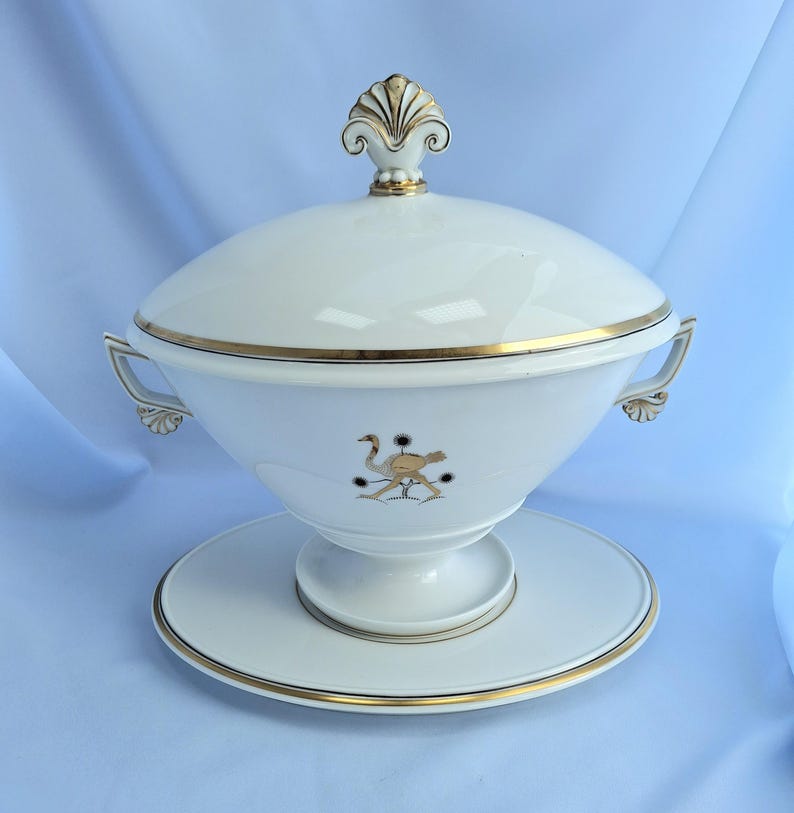 May include: A white ceramic soup tureen with a lid, handles, and a matching base. The tureen is decorated with gold trim and a stylized ostrich design. The lid is topped with a gold finial.