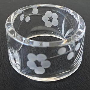 May include: A clear glass napkin ring featuring a delicate etched floral pattern with five-petaled blossoms and small circular buds. The cylindrical band is transparent with a smooth, polished finish, showcasing the frosted white etched details.