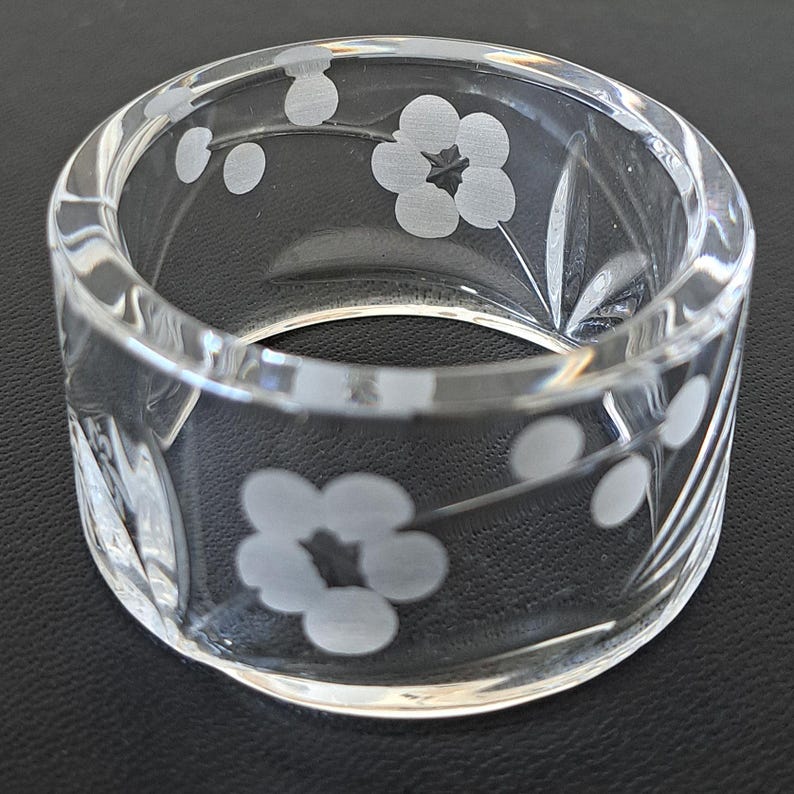 May include: A clear glass napkin ring featuring a delicate etched floral pattern with five-petaled blossoms and small circular buds. The cylindrical band is transparent with a smooth, polished finish, showcasing the frosted white etched details.