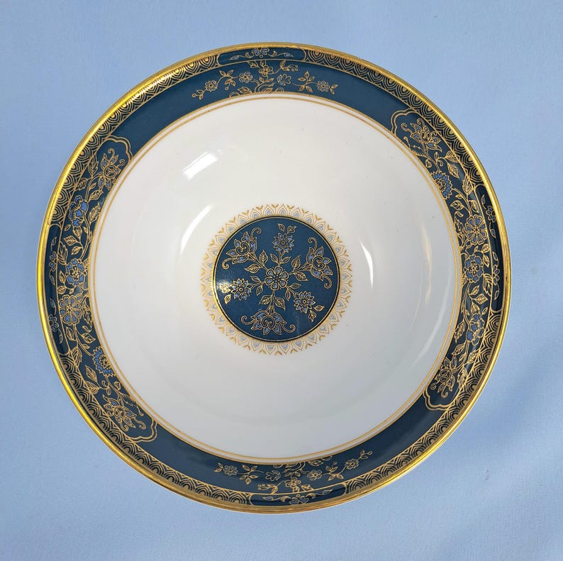 Op de afbeelding: Een ronde keramische kom met een wit midden en een cirkelvormig blauw en goudkleurig bloemenmedaillon. De brede rand is diepblauw met een goudkleurig bloemmotief en een dunne goudkleurige rand. Het binnenoppervlak is glad en glanzend.