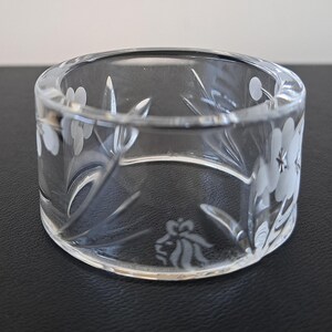 May include: A clear glass napkin ring featuring a delicate etched floral pattern with white frosted petals and leaves. The cylindrical band has a smooth, polished finish and a circular opening, showcasing a detailed botanical design on the exterior.