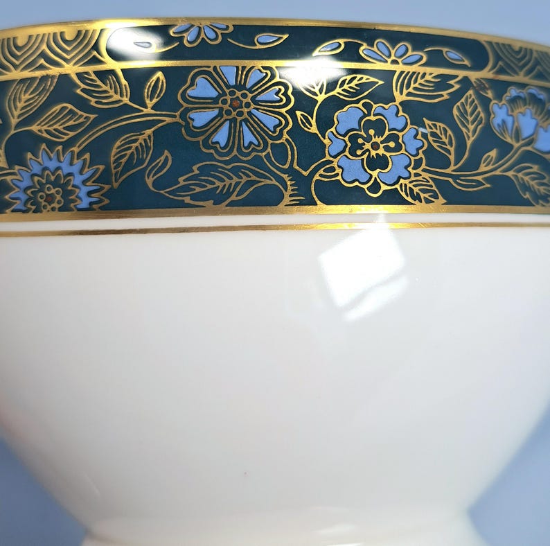 May include: A white porcelain bowl featuring a wide decorative band around the upper rim. The band is dark teal with a repeating pattern of light blue flowers and gold-colored leaves, accented by thin gold lines bordering the design.