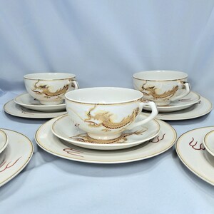 May include: White teacups and saucers featuring gold dragon motifs. The cups have handles and sit on matching saucers with gold edging. The dragon design is detailed and covers the cup and saucer. The set is on a light blue background.