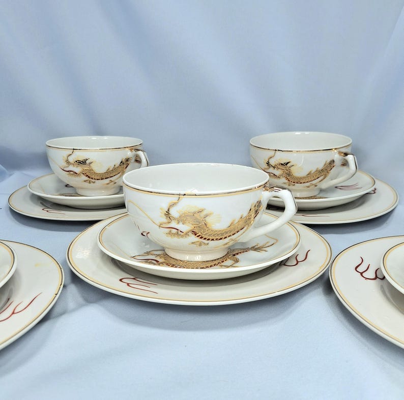 May include: White teacups and saucers featuring gold dragon motifs. The cups have handles and sit on matching saucers with gold edging. The dragon design is detailed and covers the cup and saucer. The set is on a light blue background.