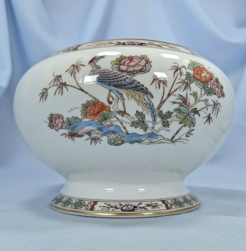Puede incluir: Un cuenco decorativo de cer&aacute;mica blanca con una base ancha y un cuerpo redondeado. El cuenco presenta una colorida ilustraci&oacute;n de un p&aacute;jaro, flores y follaje. El borde y la base est&aacute;n decorados con un patr&oacute;n marr&oacute;n y dorado.
