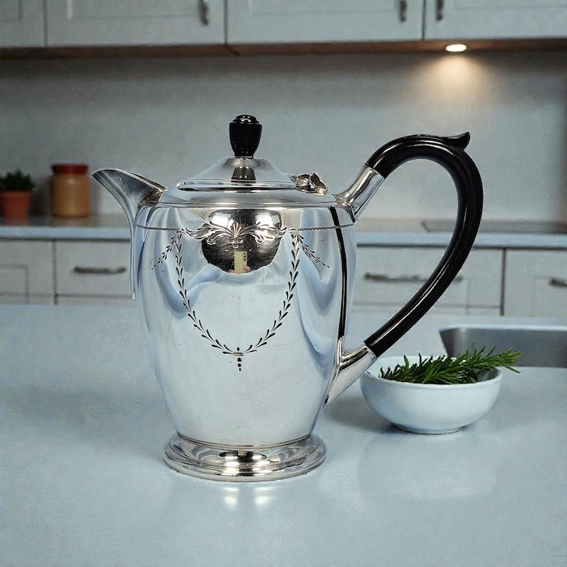 K&ouml;nnte beinhalten: Eine wei&szlig;e Keramik-Tasse mit schwarzem Henkel und dem Text "But First, Coffee" in handschriftlichem Stil. Die Tasse steht auf einer wei&szlig;en Oberfl&auml;che. Die Tasse ist etwa 10 cm hoch.