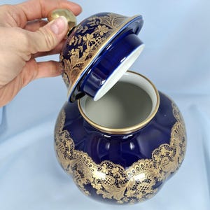 Może przedstawiać: Głęboko niebieski ceramiczny słoik z zdejmowaną pokrywką, ozdobiony misternym, złotym wzorem przypominającym koronkę. Słoik ma zaokrąglony, pękaty kształt ze złotym wykończeniem krawędzi oraz złotym uchwytem na szczycie pokrywki.