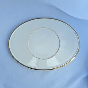 May include: An oval, off-white plate with a gold rim and a gold inner circle. The plate is set against a light blue background. This elegant dish is suitable for serving appetizers or desserts.