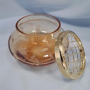 May include: A round, amber-colored glass bowl featuring a delicate etched leaf and vine pattern around its exterior. Beside the bowl sits a circular, gold-toned metal grid lid designed to fit over the opening, providing a structured top.