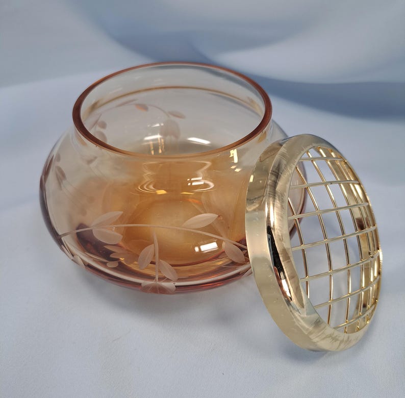 May include: A round, amber-colored glass bowl featuring a delicate etched leaf and vine pattern around its exterior. Beside the bowl sits a circular, gold-toned metal grid lid designed to fit over the opening, providing a structured top.