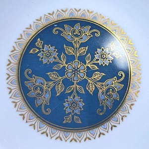 Op de afbeelding: Een ronde keramische bord met een diepblauw centrum versierd met een symmetrisch bloemmotief in goud en lichtblauw. Het ontwerp bevat een centrale bloem omringd door vier uitstralende bloesems en bladeren, omlijst door een gouden bloemblaadjesrand.