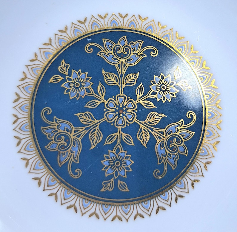 Op de afbeelding: Een ronde keramische bord met een diepblauw centrum versierd met een symmetrisch bloemmotief in goud en lichtblauw. Het ontwerp bevat een centrale bloem omringd door vier uitstralende bloesems en bladeren, omlijst door een gouden bloemblaadjesrand.