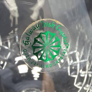 May include: A clear lead crystal object featuring a circular sticker with green text and a green starburst graphic. The text reads Galway Irish Crystal and OVER 24% LEAD CRYSTAL. The surface of the crystal displays a faceted, geometric pattern.