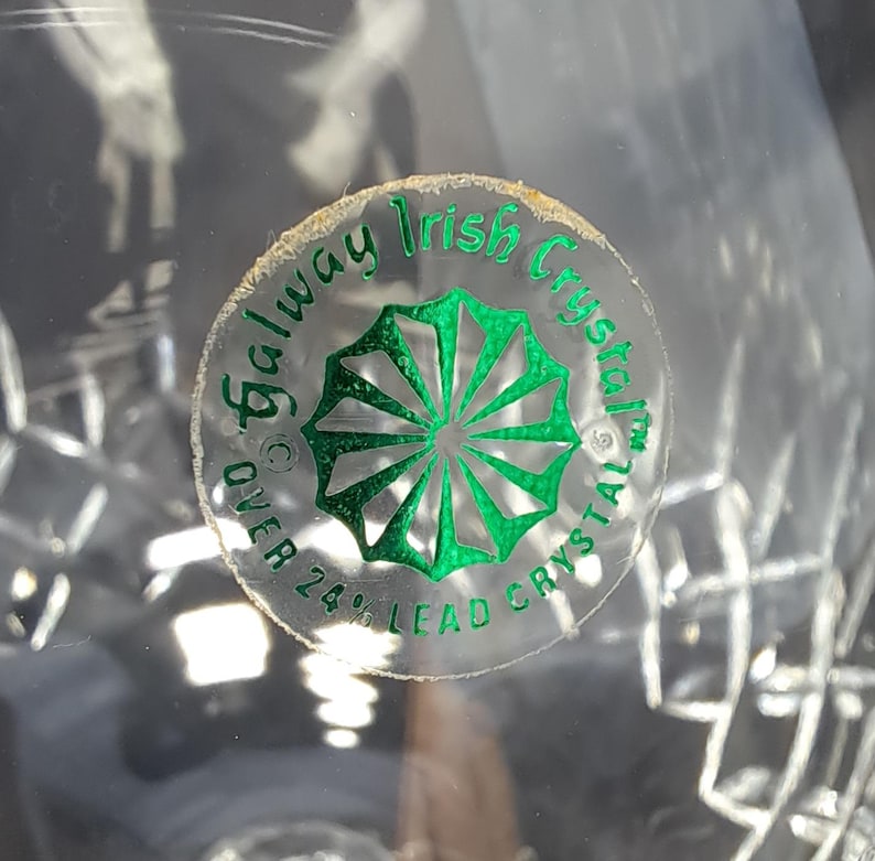 May include: A clear lead crystal object featuring a circular sticker with green text and a green starburst graphic. The text reads Galway Irish Crystal and OVER 24% LEAD CRYSTAL. The surface of the crystal displays a faceted, geometric pattern.