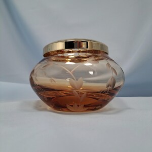 May include: A round, amber-colored glass jar featuring a delicate etched floral pattern on its exterior. The jar is topped with a polished, reflective gold-tone metal lid that sits securely on the rim, creating a smooth and cohesive appearance.