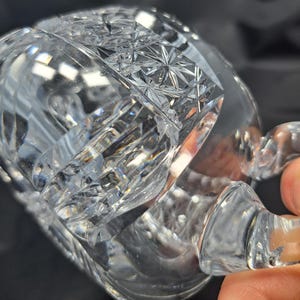 Op de afbeelding: Een heldere glazen beker met ingewikkelde geometrische slijppatronen over het oppervlak. Het ontwerp bevat een opvallende band met stermotieven en verticale groeven, met een gebogen handvat aan de zijkant van de beker.