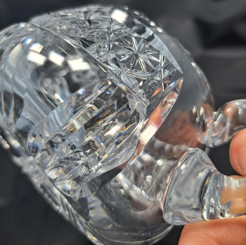 Op de afbeelding: Een heldere glazen beker met ingewikkelde geometrische slijppatronen over het oppervlak. Het ontwerp bevat een opvallende band met stermotieven en verticale groeven, met een gebogen handvat aan de zijkant van de beker.