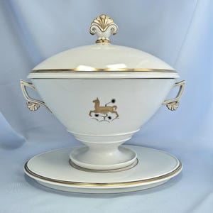 May include: A white porcelain soup tureen with a lid, gold trim, and a matching plate. The tureen features a gold emblem of a deer and floral accents. The lid has a decorative gold finial. The handles are also gold.