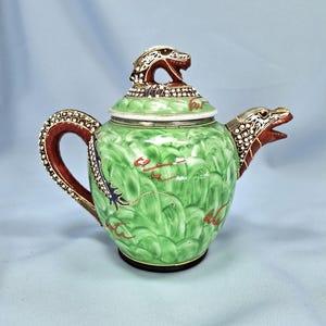Pu&ograve; includere: Una teiera in ceramica verde con un design a drago. La teiera presenta una testa di drago sul coperchio e sul beccuccio, con un manico rosso e dorato. Il corpo ha un motivo strutturato con accenti rossi e dorati.