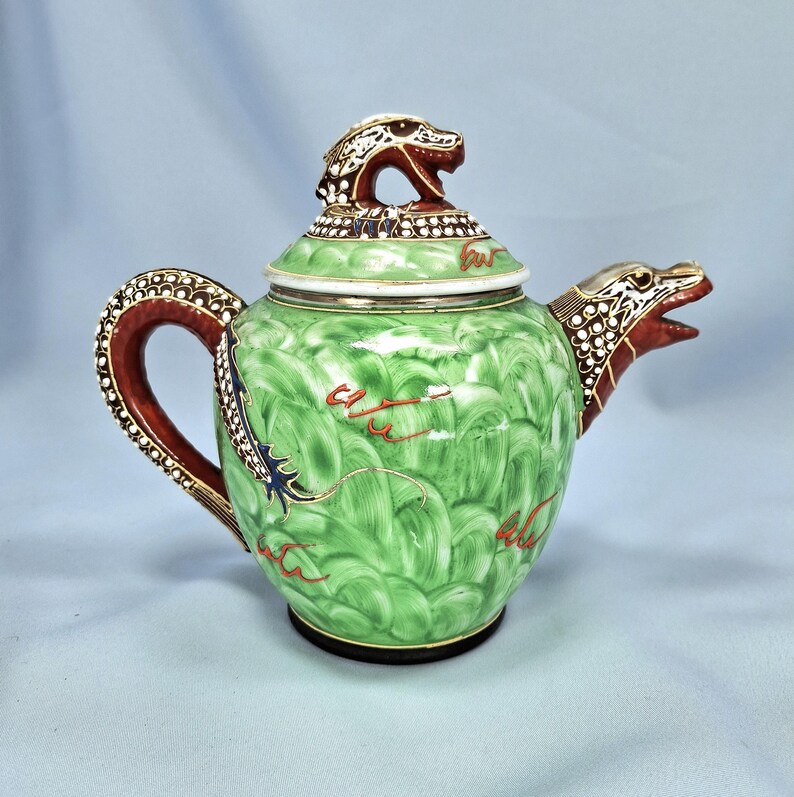 Pu&ograve; includere: Una teiera in ceramica verde con un design a drago. La teiera presenta una testa di drago sul coperchio e sul beccuccio, con un manico rosso e dorato. Il corpo ha un motivo strutturato con accenti rossi e dorati.