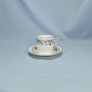 Peut inclure: Un ensemble tasse et soucoupe blanc avec un motif floral. La tasse a une anse et un bord dor&eacute;s, et la soucoupe a un bord dor&eacute; assorti. Le motif floral comprend des fleurs roses, jaunes et vertes.