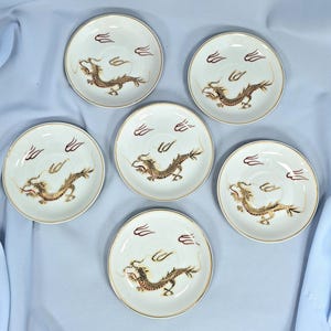 May include: Six white ceramic saucers with a gold rim and a detailed gold and brown dragon design. Each saucer has a stylized cloud design in maroon. A classic Asian-inspired design.
