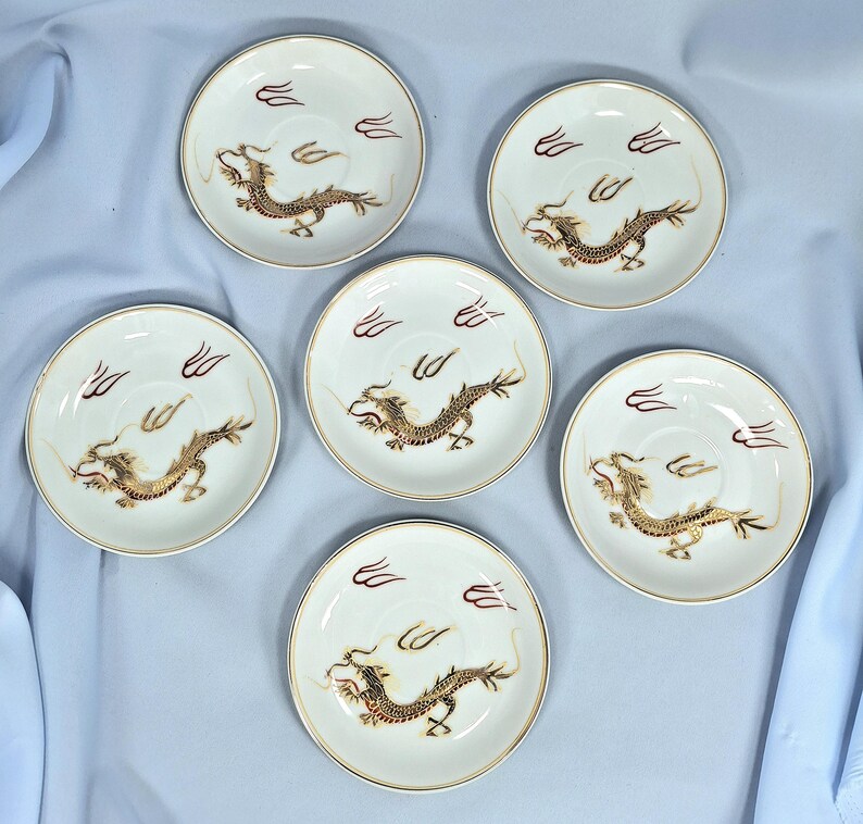 May include: Six white ceramic saucers with a gold rim and a detailed gold and brown dragon design. Each saucer has a stylized cloud design in maroon. A classic Asian-inspired design.