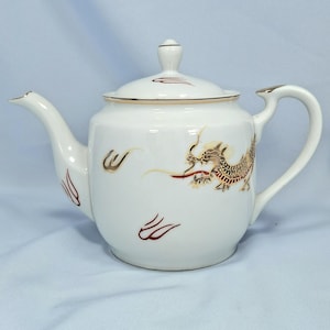 May include: White ceramic teapot with a lid and a curved handle. The teapot is decorated with a gold and brown dragon design on the side. Gold trim accents the lid and handle. Red and gold accents are also present.