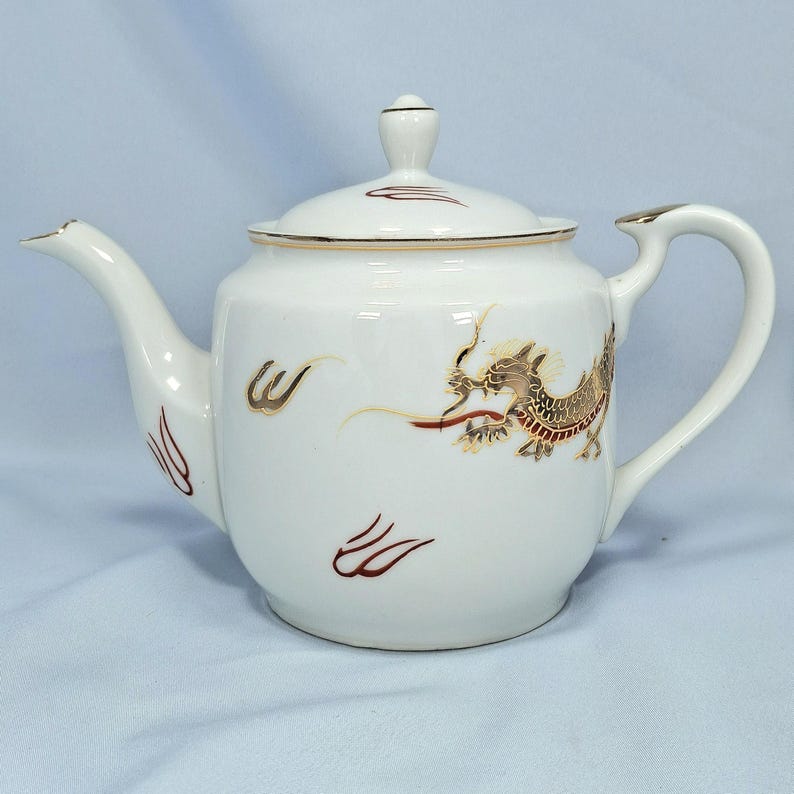 May include: White ceramic teapot with a lid and a curved handle. The teapot is decorated with a gold and brown dragon design on the side. Gold trim accents the lid and handle. Red and gold accents are also present.