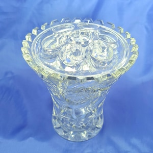 May include: A clear cut glass flower frog vase featuring a removable top insert with seven circular openings. The body of the vase displays a faceted geometric pattern with a serrated rim, reflecting light through its transparent glass structure.