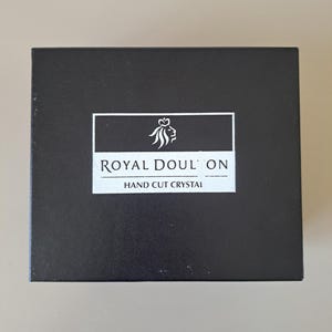 May include: A rectangular black box featuring a white rectangular label in the centre. The label displays a white lion head logo above the text ROYAL DOULTON and the words HAND CUT CRYSTAL printed in white capital letters on a black background.