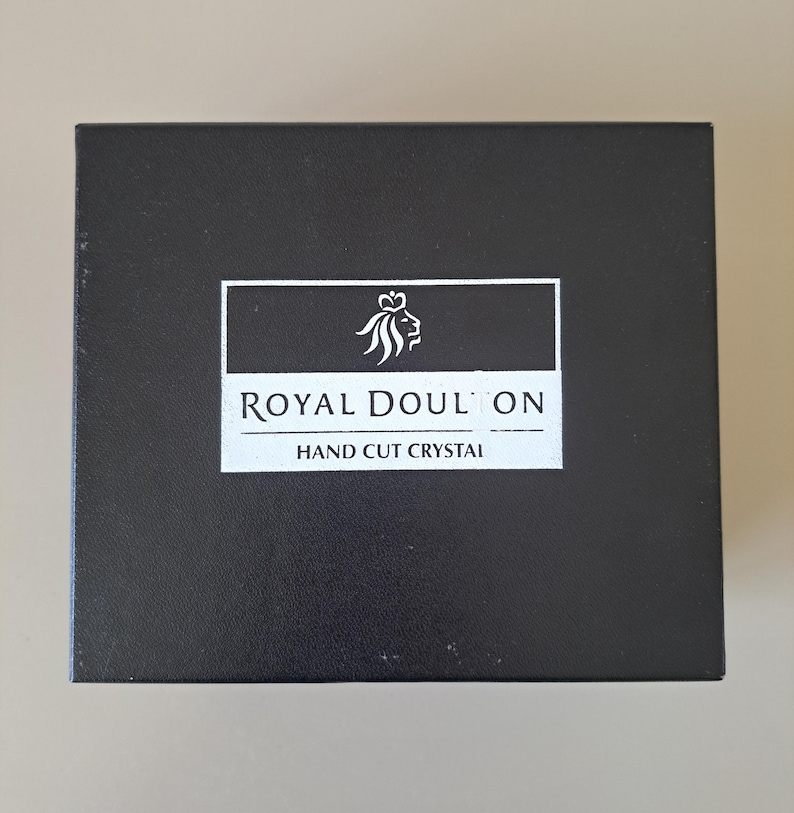 May include: A rectangular black box featuring a white rectangular label in the centre. The label displays a white lion head logo above the text ROYAL DOULTON and the words HAND CUT CRYSTAL printed in white capital letters on a black background.